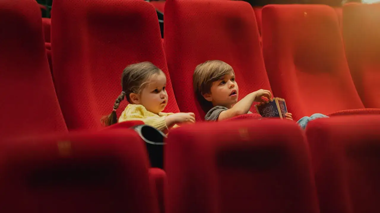 Två barn sitter i röda biosäten och tittar på filmer på bio nu. En av dem håller en popcornpåse.