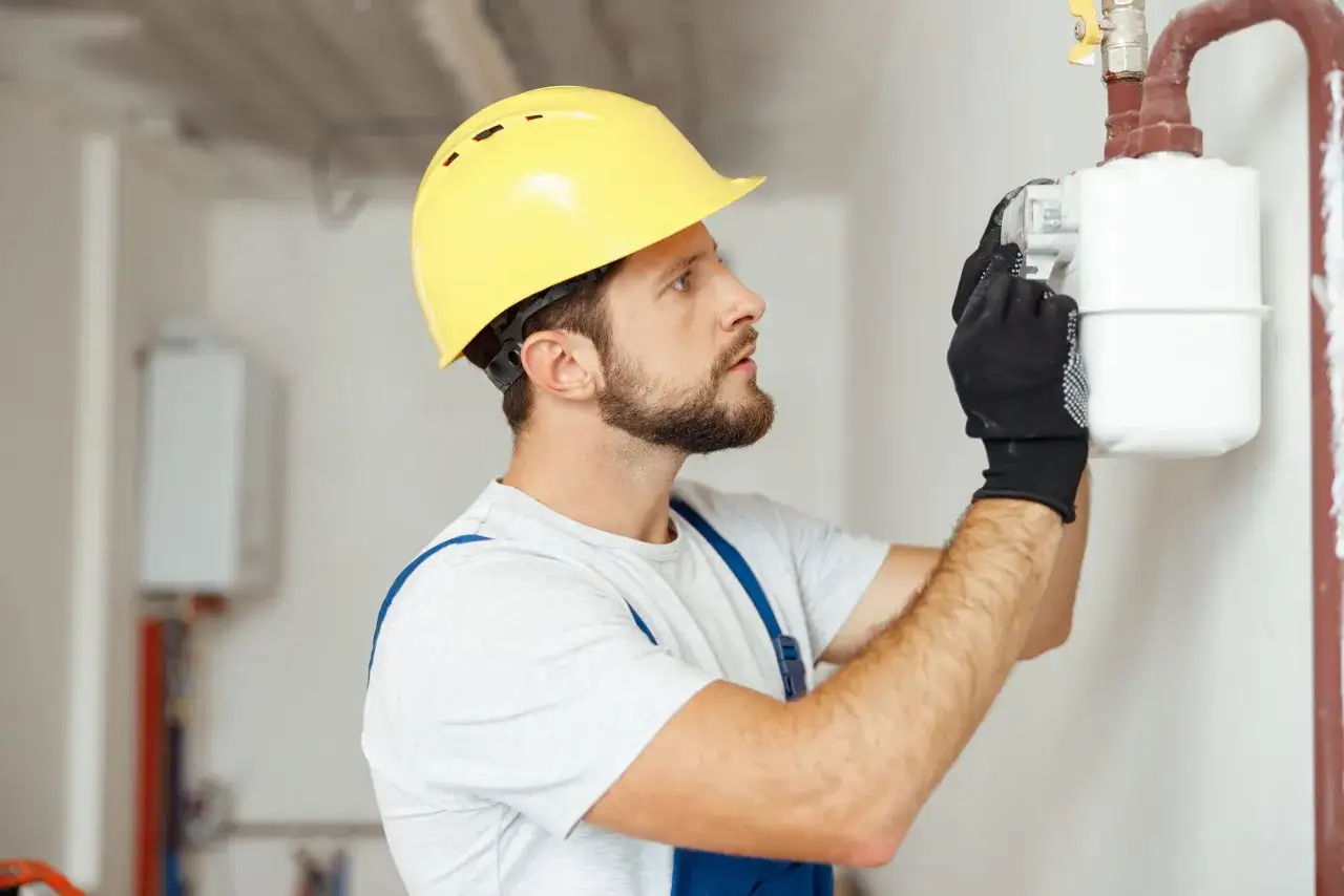 Instalator w żółtym kasku sprawdza licznik gazowy, pokazując, jak sprawdzić szczelność gazu w domu.