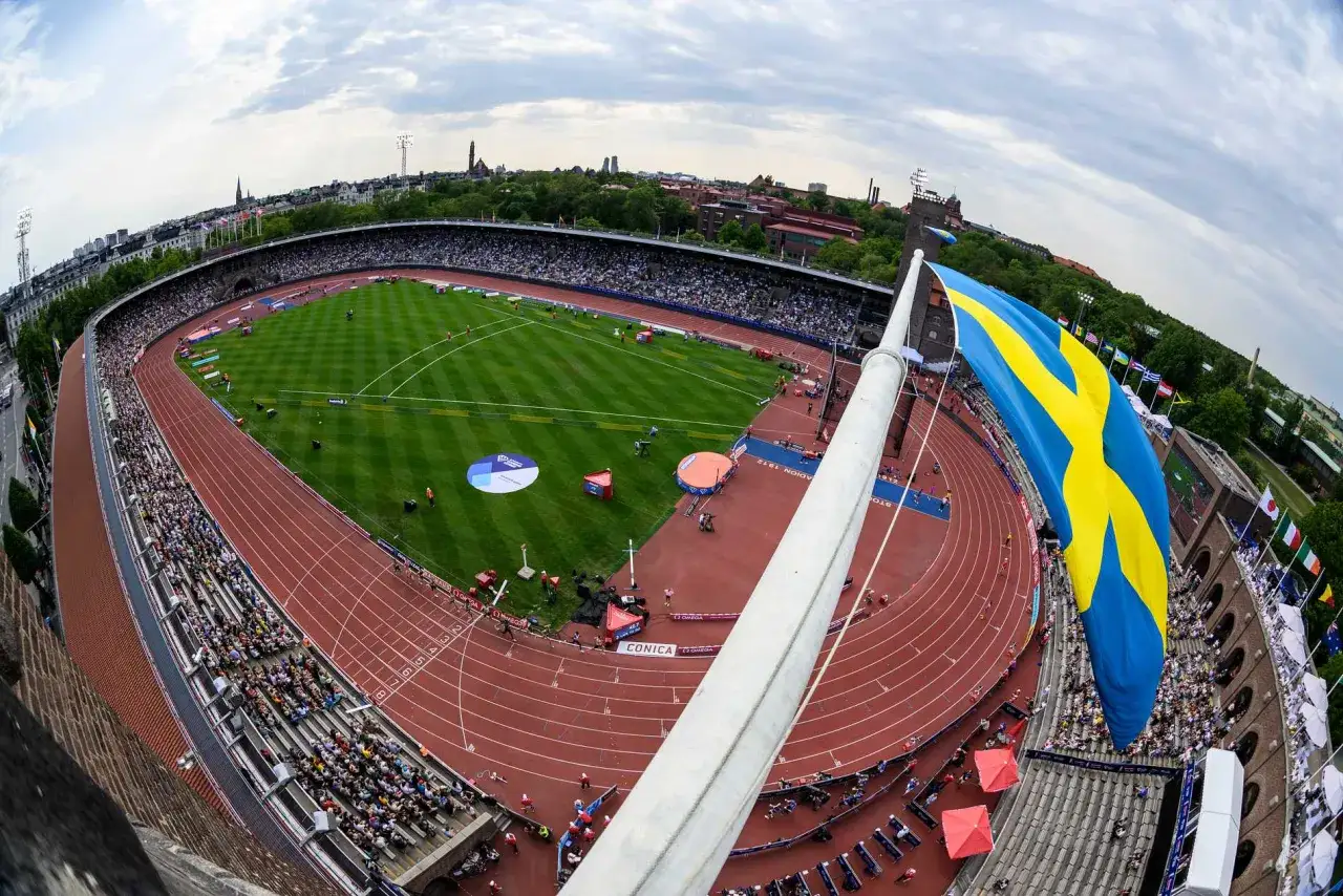 Stor svensk flagga vajar över en fullsatt arena i Stockholm. En friidrottstävling pågår på den röda löparbanan och gröna gräsmattan.