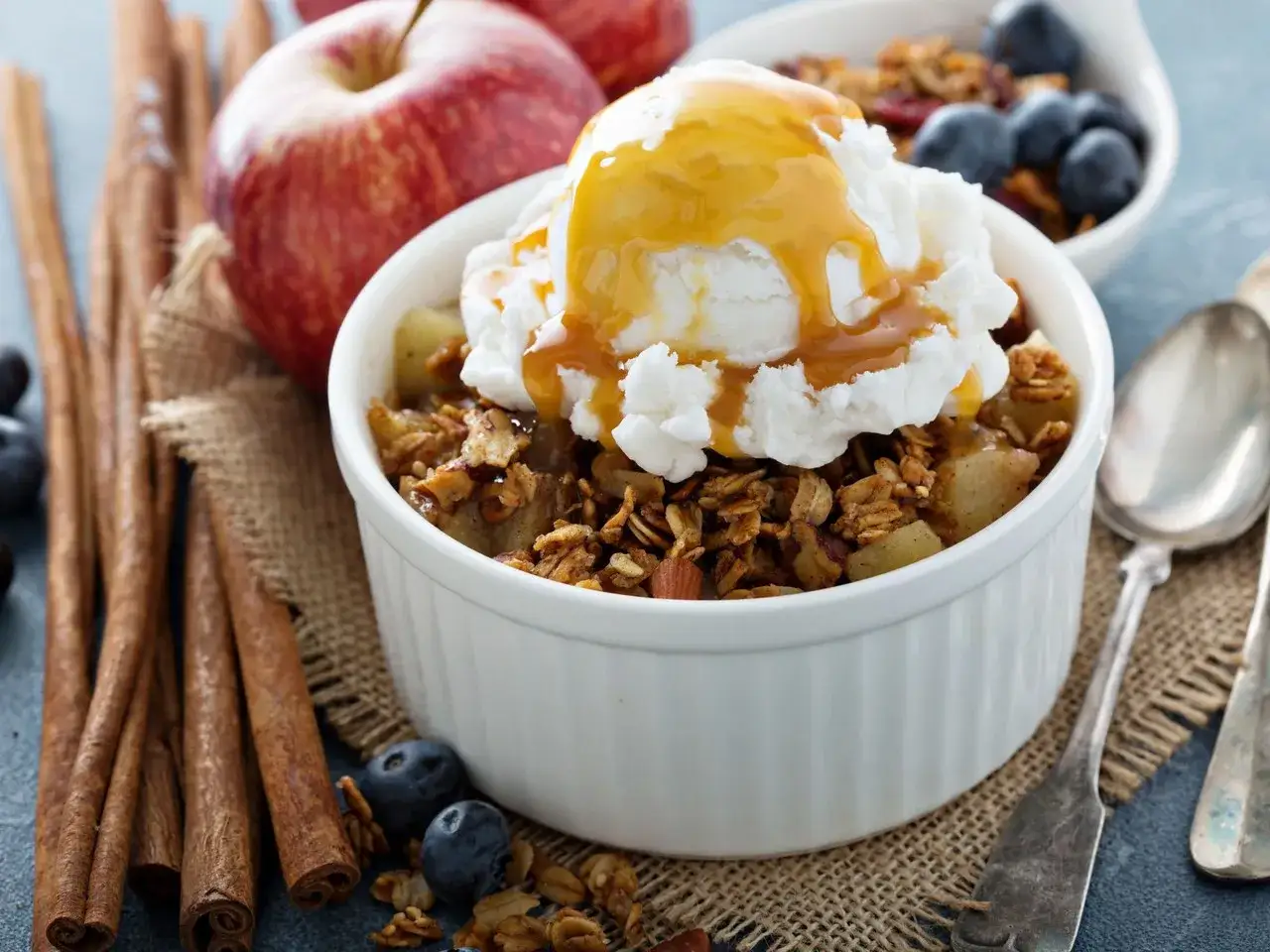 Delicioso crumble de manzana con helado y caramelo, rodeado de canela y arándanos.