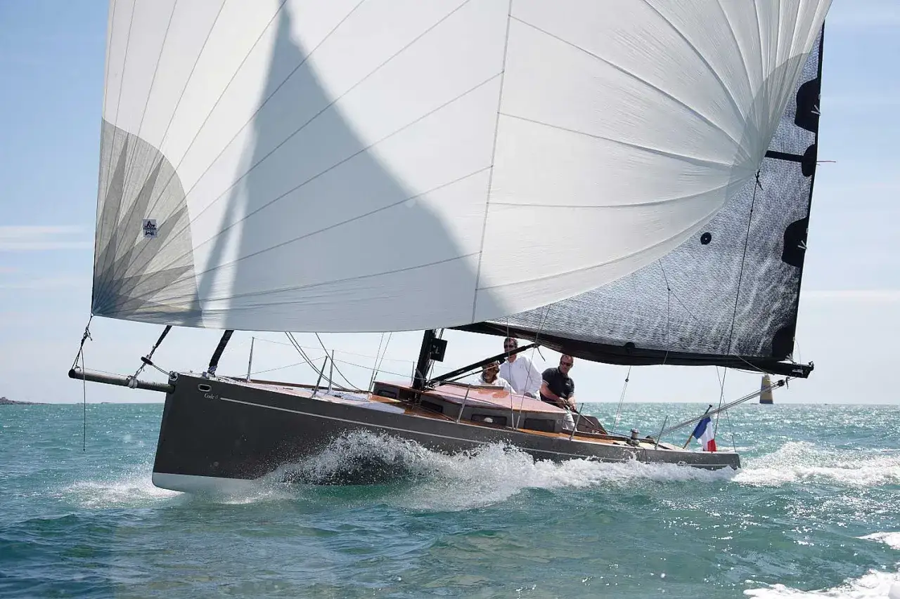 Barca a vela con equipaggio che naviga con vento forte. La grande vela bianca, un code 0, cattura il vento, mentre l'onda si infrange sullo scafo.