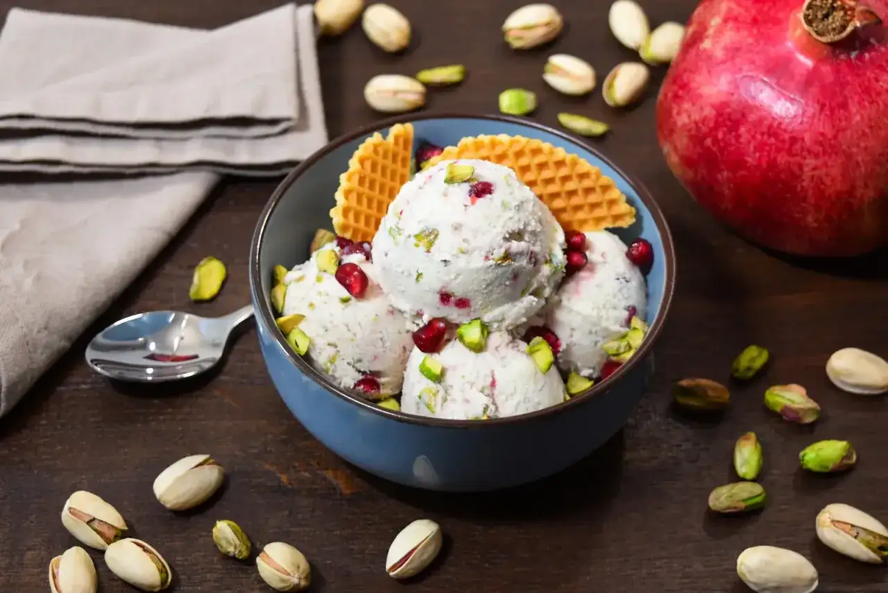 Sch&uuml;ssel mit cremigem Granatapfel Eis, garniert mit Granatapfelkernen und Pistazien. Daneben liegen ganze Granat&auml;pfel und Pistazien.