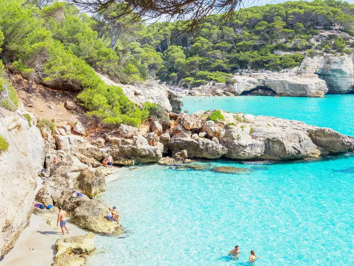 Spiaggia spagnola con acque cristalline, scogliere rocciose e vegetazione rigogliosa. Perfetta per le mete estive in Spagna, con persone che nuotano e si rilassano.