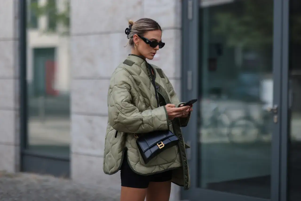 Kobieta w stylowej, pikowanej kurtce wiosennej khaki, z torebką na ramieniu i telefonem w dłoni, gotowa na miejskie wyzwania.