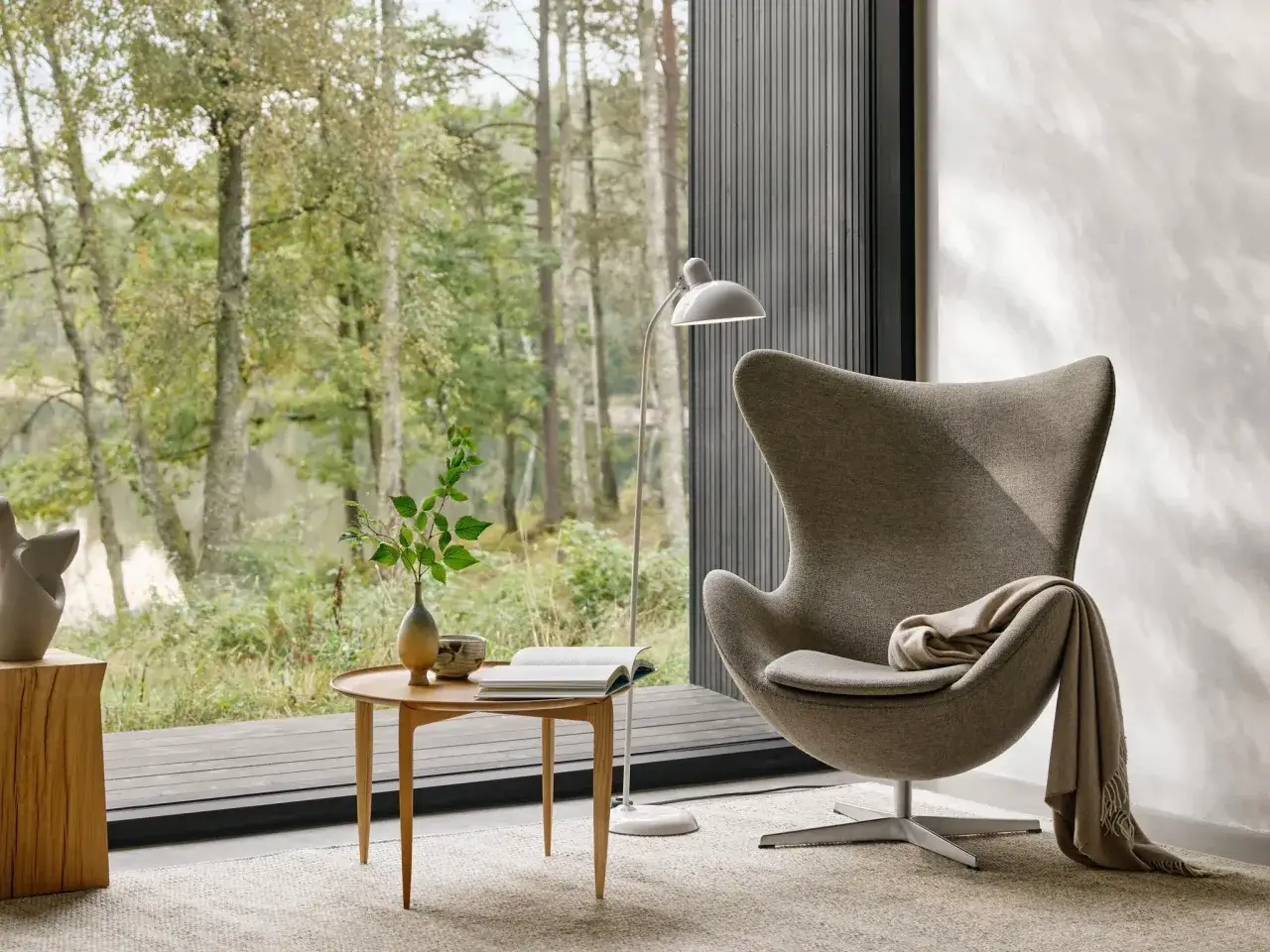 Ein Sessel wird mit einer Decke dekoriert. Daneben ein Beistelltisch mit Buch und Vase. Gro&szlig;e Fensterfront mit Blick in den Wald.