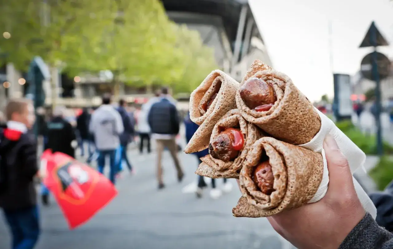 Une main tient plusieurs galettes saucisses, garniture de saucisses et de sauce, devant une foule floue près d'un stade.