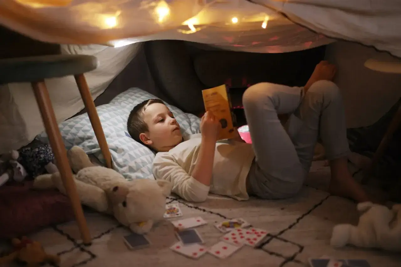 Junge liest in einer gem&uuml;tlichen H&ouml;hle, umgeben von Spielzeug und Lichterketten. Vielleicht sucht er nach Inspiration f&uuml;r neue kinder schlaflieder.