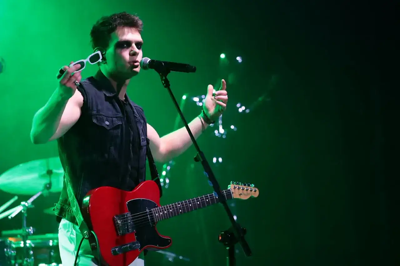 Krzysztof Cugowski Junior na scenie z czerwoną gitarą i okularami w dłoni, w otoczeniu zielonego światła.