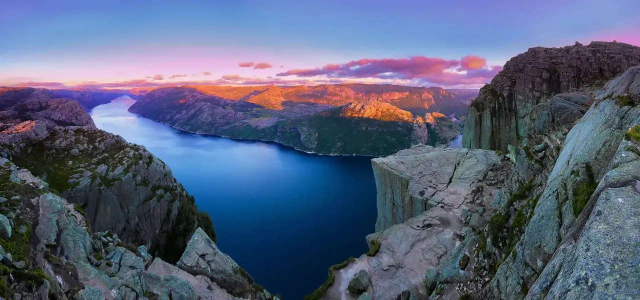 Solnedgång över Stavangerfjorden, med Preikestolen som sticker ut. Dramatiska klippor möter det djupblå vattnet under en himmel i rosa och lila toner.