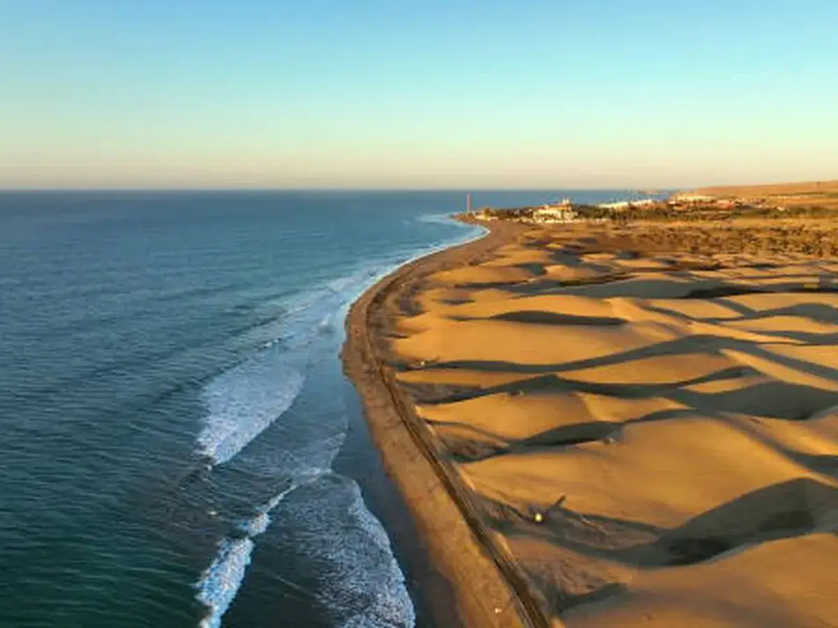 Oceano azul y dunas doradas en las **playas de Canarias**. Un faro y edificios se vislumbran al fondo.