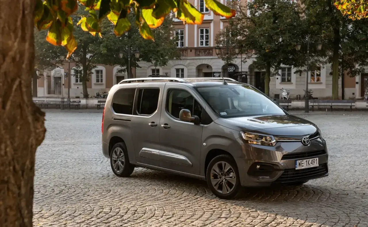 Szary Toyota Proace City Verso używane na brukowanym placu, z drzewami i budynkami w tle. Idealny do rodzinnych podr&oacute;ży.