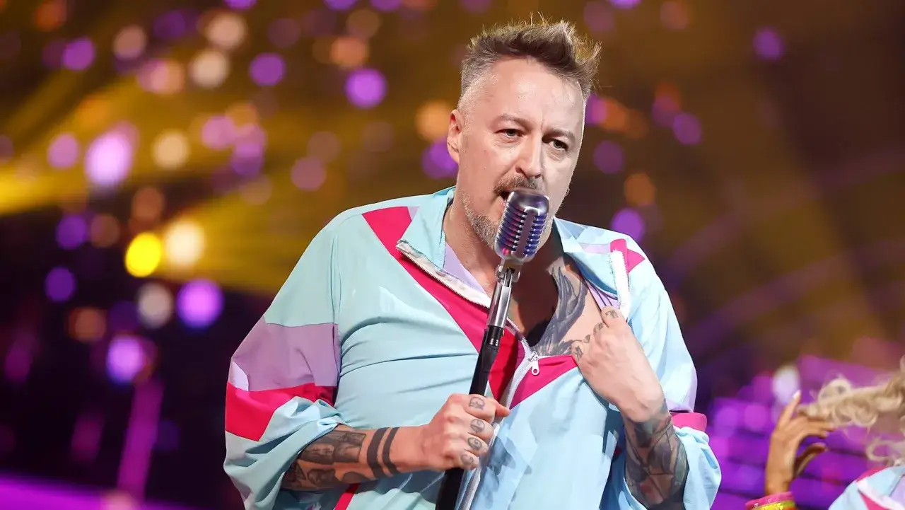 Piotr Kędzierski, z charakterystycznym tatuażem na klatce piersiowej, śpiewa do mikrofonu. Jego wzrost nie jest widoczny, ale energia sceniczna jest wyczuwalna.