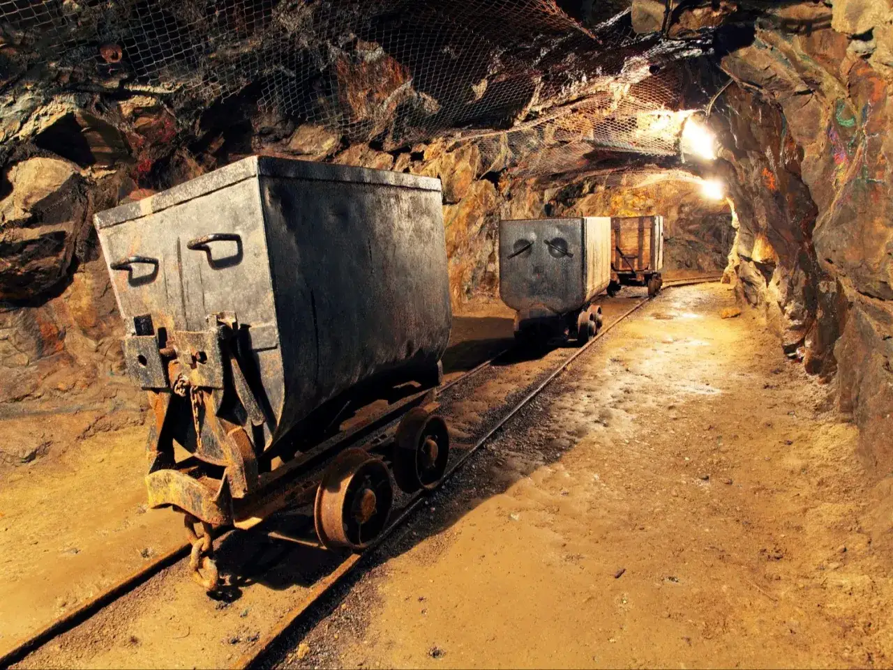 W kopalni, w tunelu oświetlonym lampami, stoją wagony górnicze. Czekają na załadunek, może na cenny, **złoty pociąg** rudy.