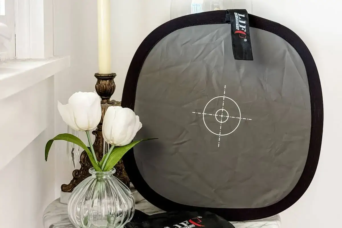 Un riflettore grigio con un mirino bianco, utile per il bilanciamento del bianco, accanto a tulipani bianchi e un candelabro.