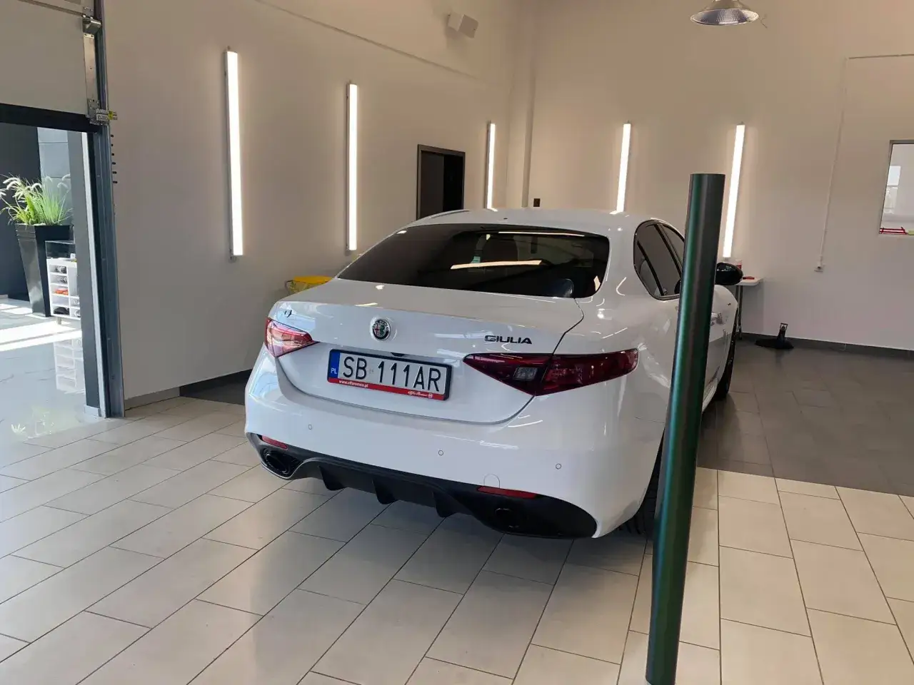 Białe Alfa Romeo Giulia w garażu, oświetlone pionowymi lampami detailingowymi.