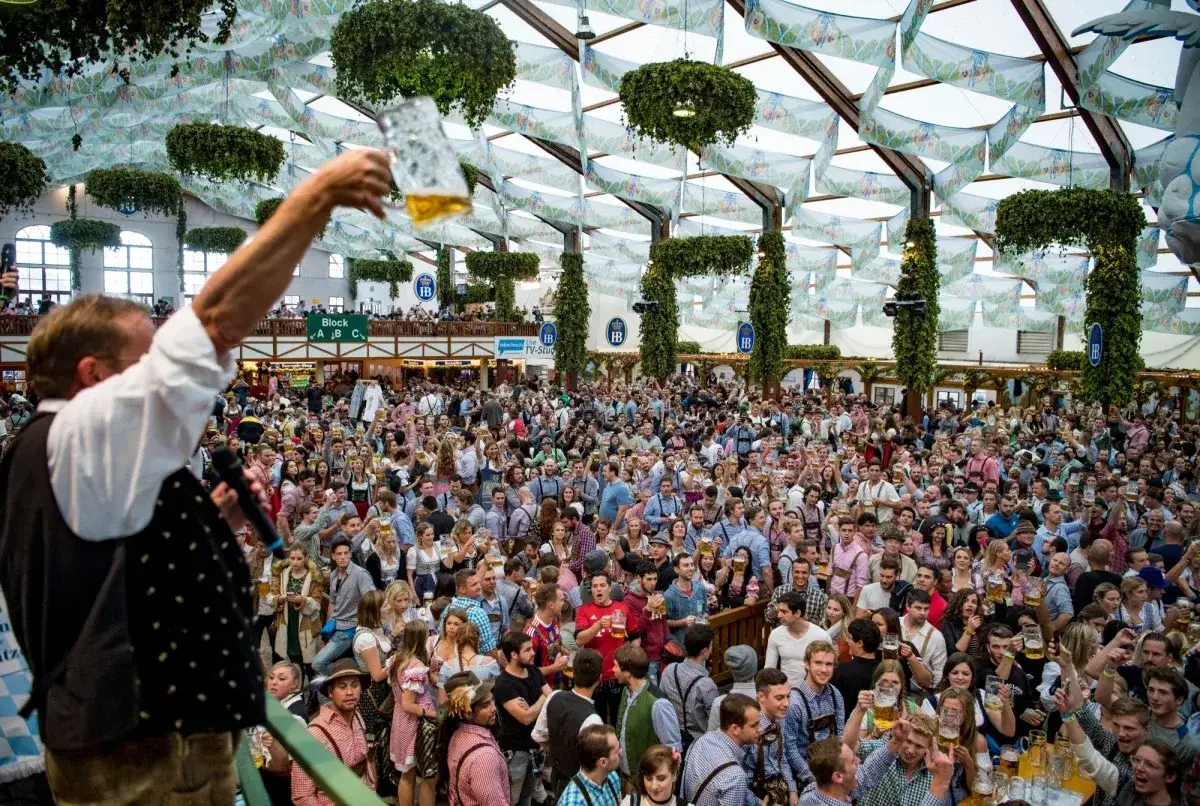 Tłum ludzi świętuje w wielkim namiocie, wznosząc kufle piwa. Widać dekoracje i scenę. To właśnie tak wygląda Oktoberfest!