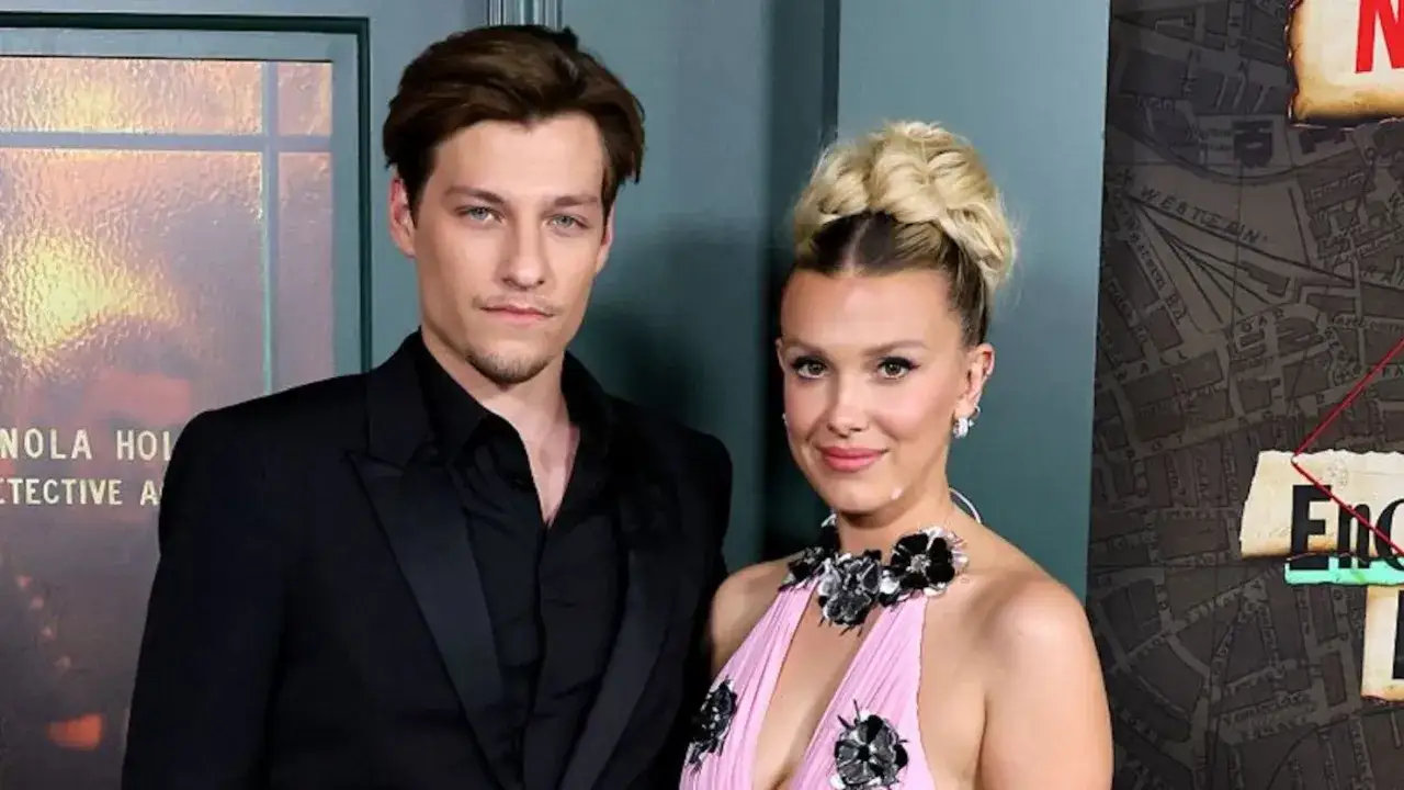 Millie Bobby Brown y su pareja posan sonrientes. Ella luce un vestido rosa con flores negras, él un traje negro.