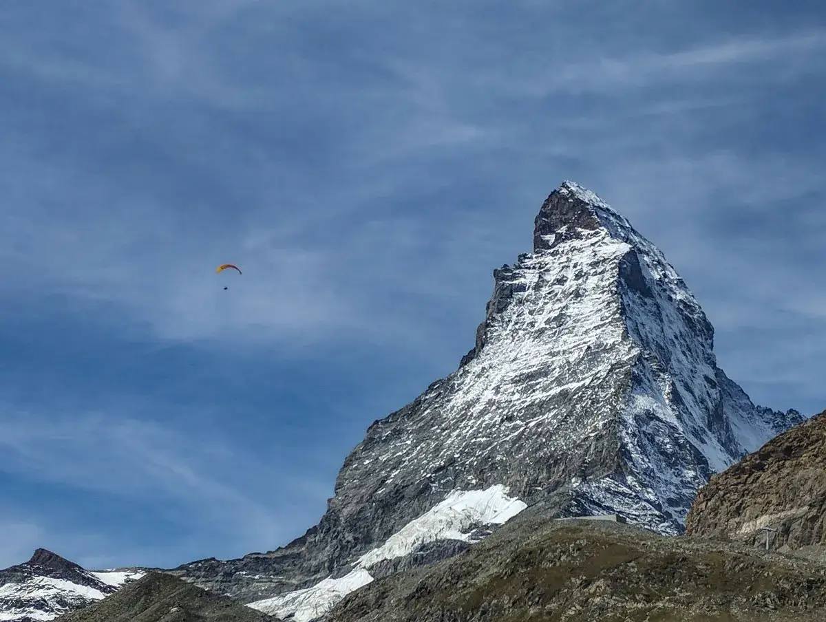 Majestatyczny Matterhorn, pokryty śniegiem, g&oacute;ruje nad krajobrazem. W oddali, na tle błękitnego nieba, szybujący paralotniarz symbolizuje wolność i przygodę, jakby przygotowywał się do wejścia na Matterhorn.