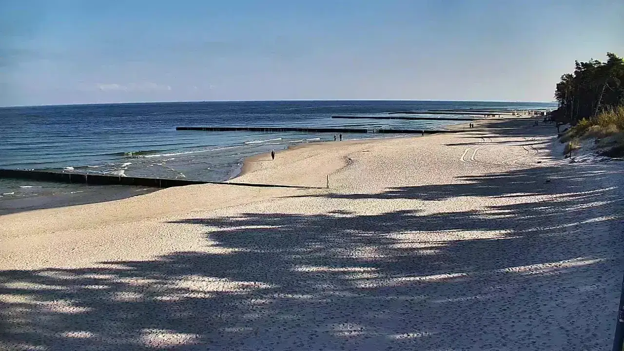Spokojna plaża w Dźwirzynie nad morzem. Delikatne fale obmywają brzeg, a drewniane falochrony przecinają wodę. Kilka osób spaceruje po piasku.