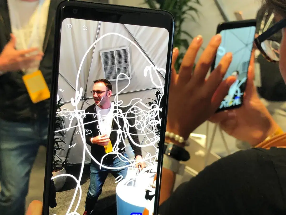 Osoba trzyma telefon, na którym widać mężczyznę z okularami i białe, chaotyczne linie. To ar doodle, czyli rysunek w rzeczywistości rozszerzonej.