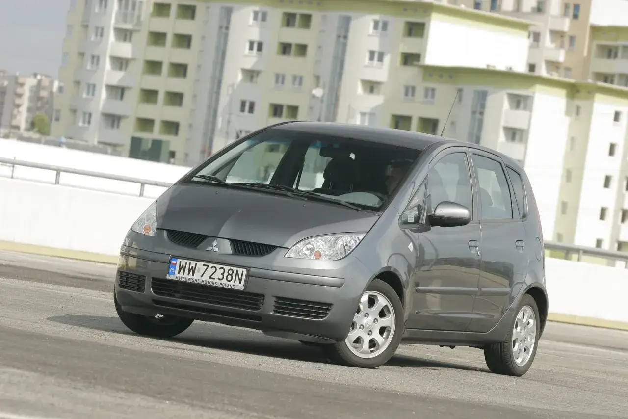 Szary Mitsubishi Colt, gotowy na mitsubishi colt wymiana żarówki przód. W tle nowoczesne budynki.