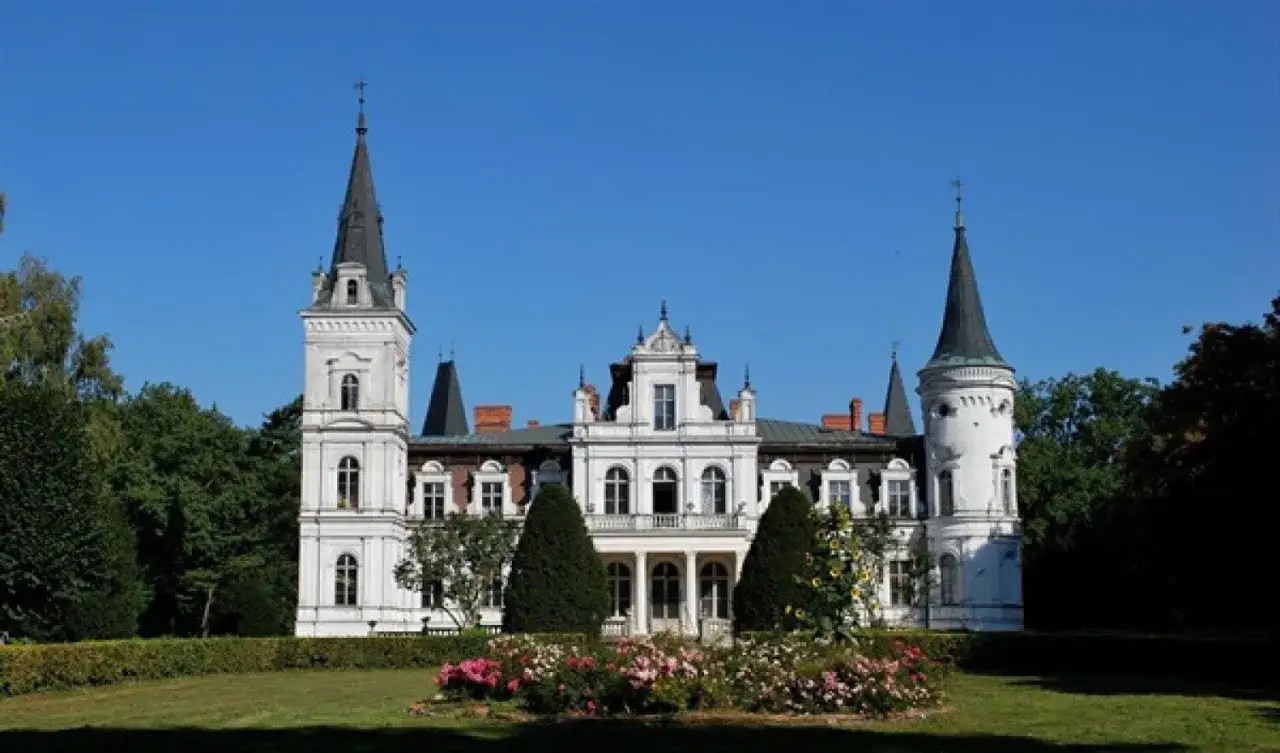 Imponujący, biały pałac z wieżami i ozdobnym ogrodem. Posadowo pałac prezentuje się majestatycznie na tle błękitnego nieba.