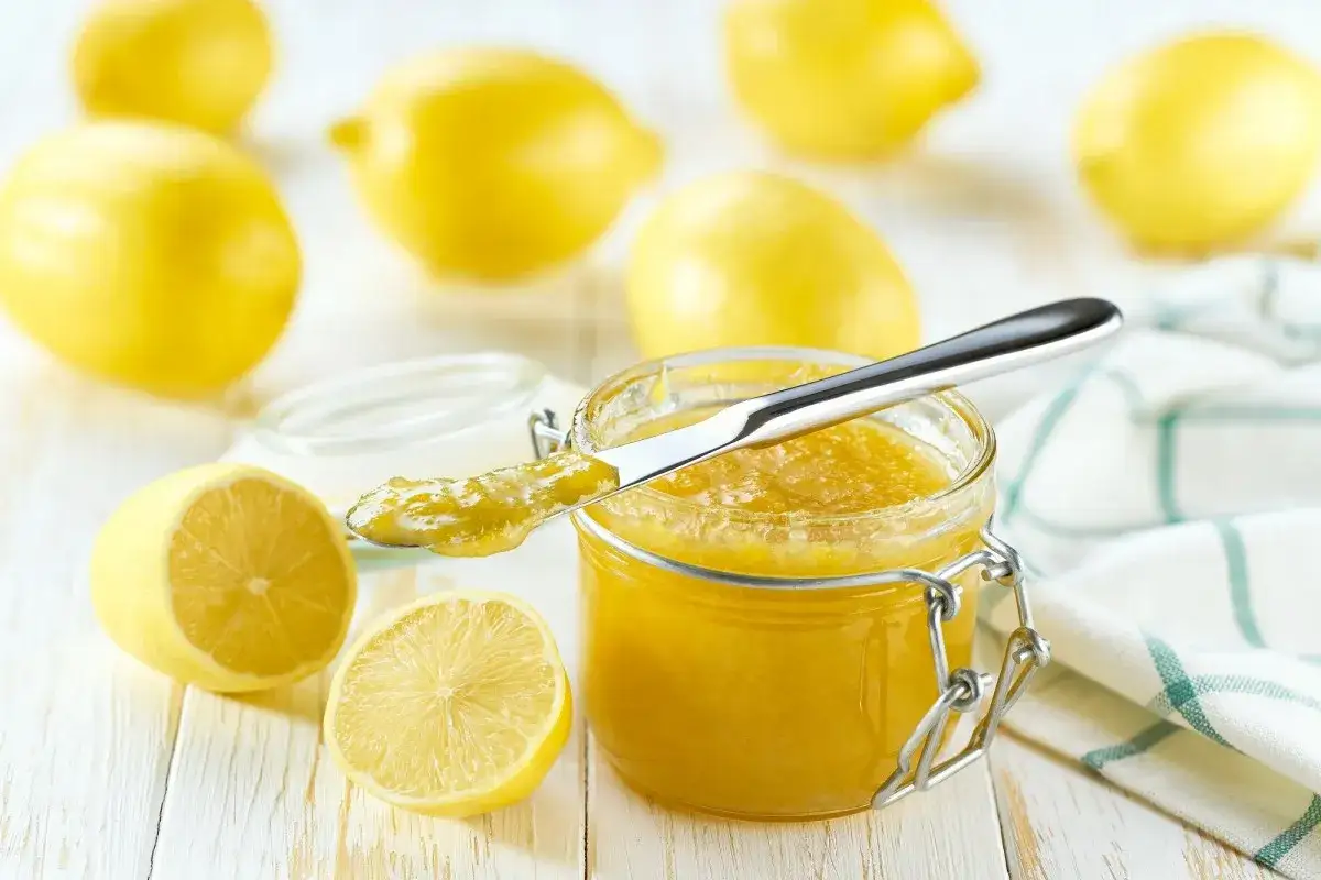 Ein Glas mit selbstgemachter Zitronenmarmelade, daneben frische Zitronen und ein Messer mit Marmelade. Perfekt für das Zitronenmarmelade Rezept!