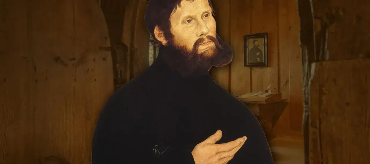 Martin Luther auf der Wartburg, im Hintergrund ein kleines Porträt von ihm und ein Schreibtisch mit Feder und Tinte.