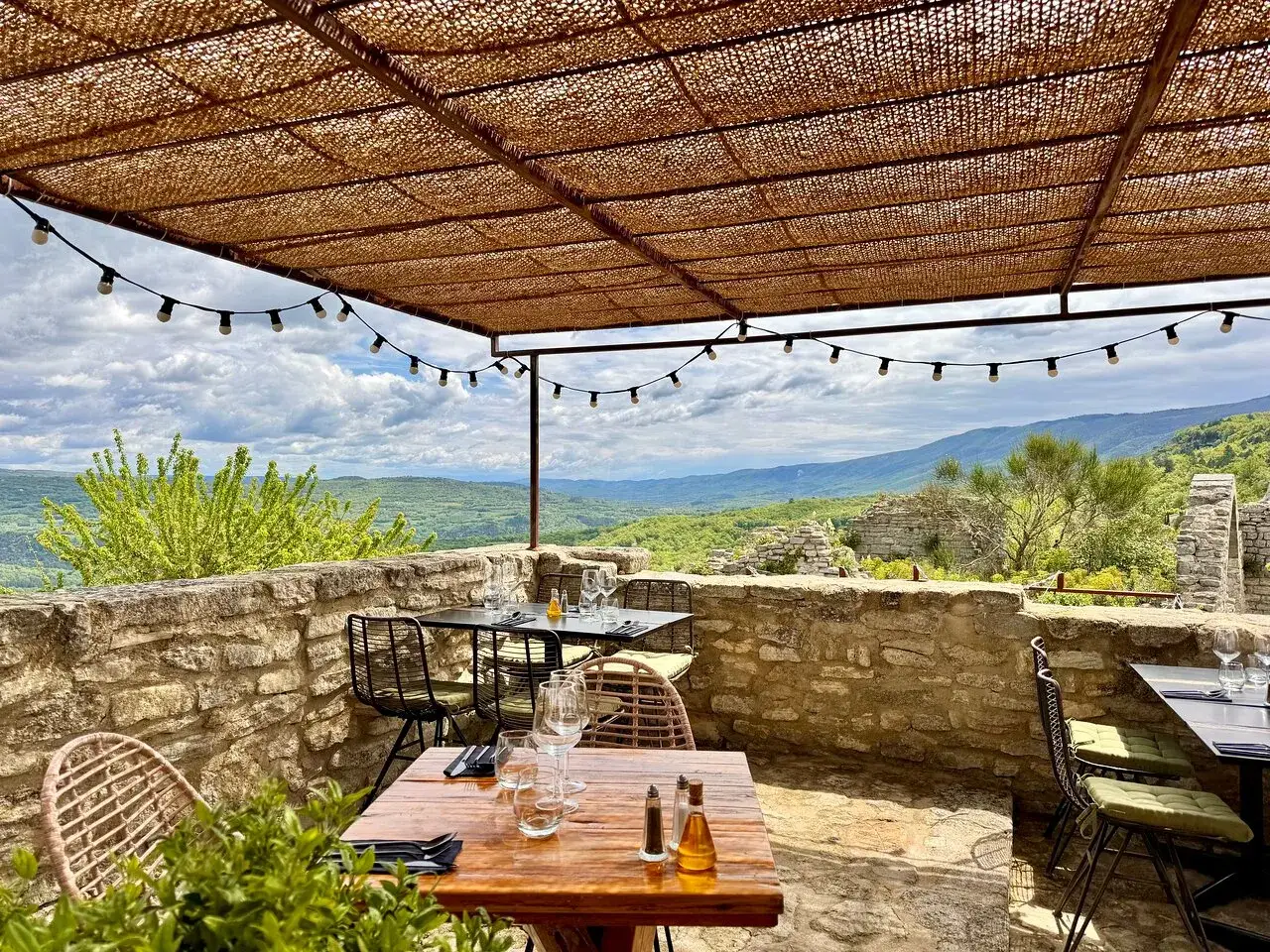 Terrasse d'un restaurant avec vue panoramique sur la vall&eacute;e. Tables dress&eacute;es sous un auvent de chaume, guirlandes lumineuses. Un jardin sur le toit Saignon invite &agrave; la d&eacute;tente.