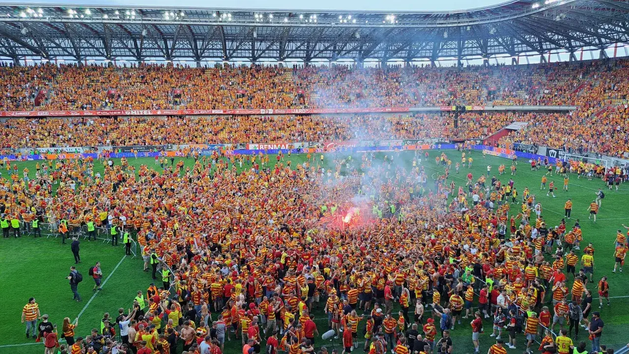 Tłum kibiców w żółto-czerwonych barwach, świętujących na murawie stadionu. Widać race i radość fanów Jagiellonii.