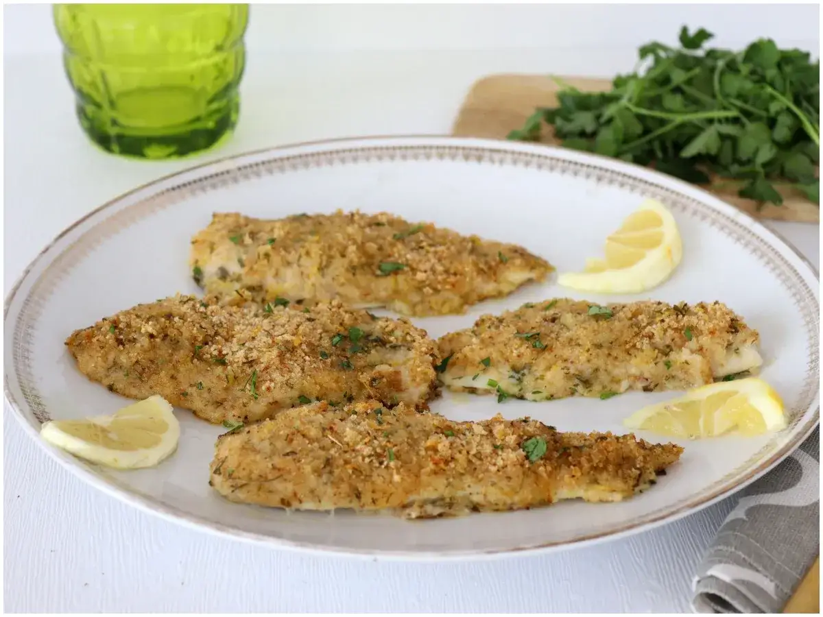 Quattro filetti di merluzzo al forno, dorati e croccanti, guarniti con prezzemolo fresco e spicchi di limone.