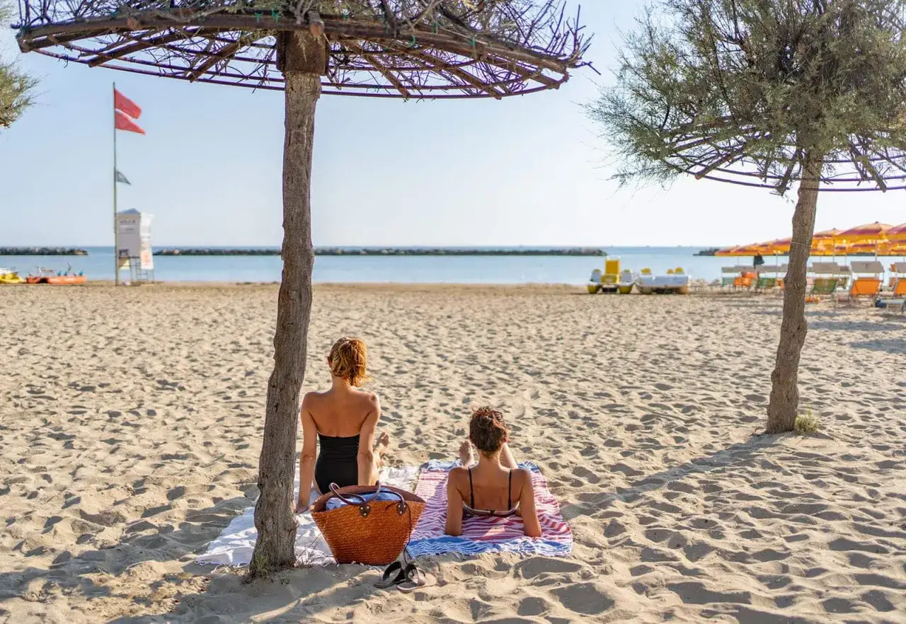 Due donne si rilassano sotto un ombrellone naturale sulla spiaggia delle tamerici, con il mare azzurro e le sdraio sullo sfondo.