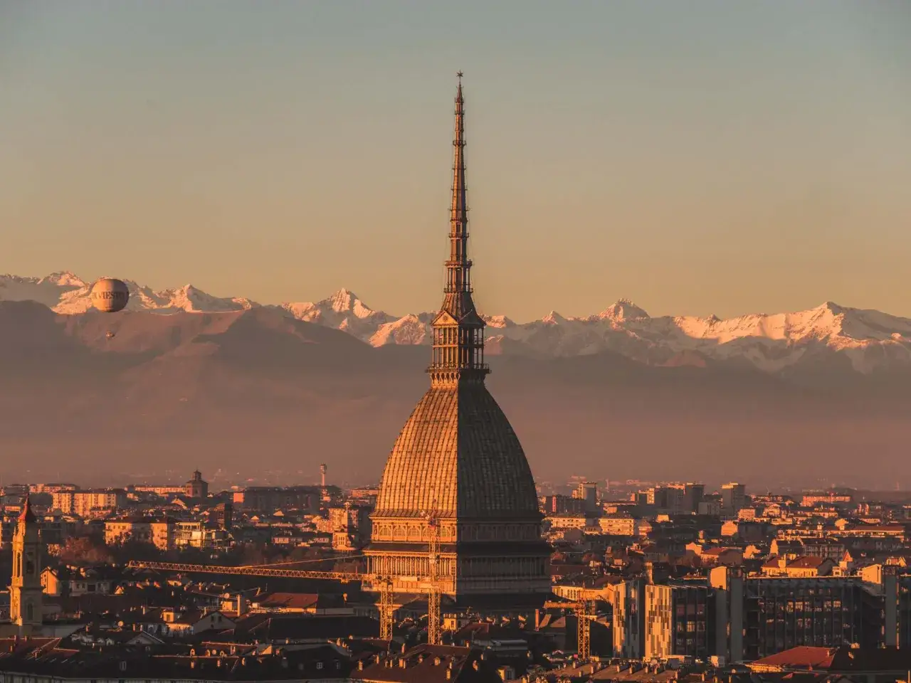 La Mole Antonelliana domina Torino al tramonto, con le Alpi innevate sullo sfondo. Un pallone aerostatico galleggia nel cielo. Perfetto per scoprire cosa vedere a Torino a piedi.