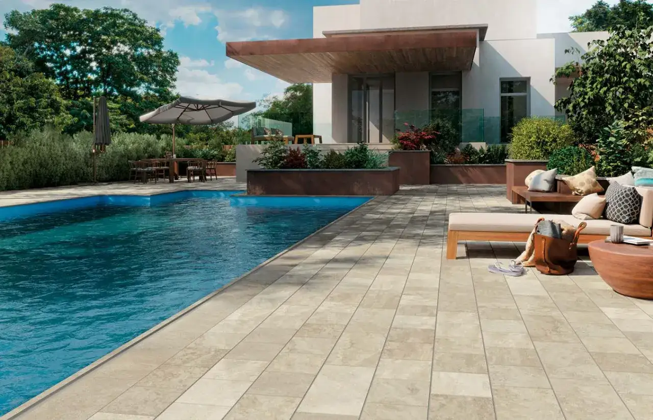 Piscina e patio con pavimentazione in pietra travertine, arredamento moderno e vegetazione lussureggiante.
