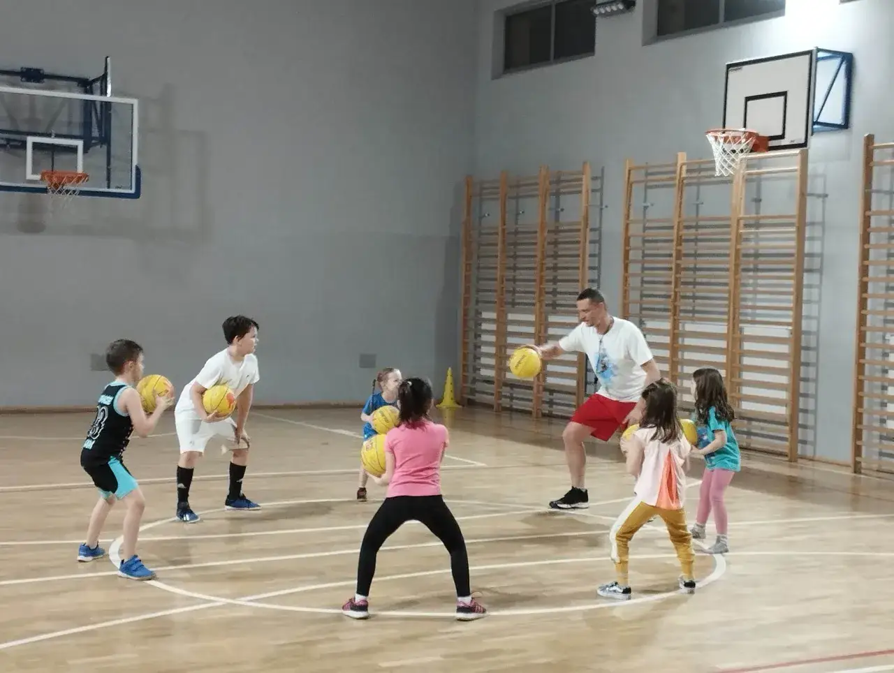 Trener prowadzi zajęcia z koszykówki dla dzieci. Mali gracze ćwiczą rzuty i podania na sali gimnastycznej.
