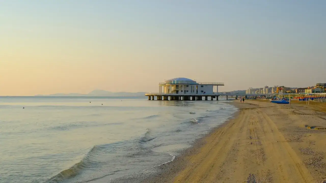 Alba dorata sul mare di Senigallia, con il Palazzetto Baviera che si staglia sull'acqua e la spiaggia che si estende verso la citt&agrave;.