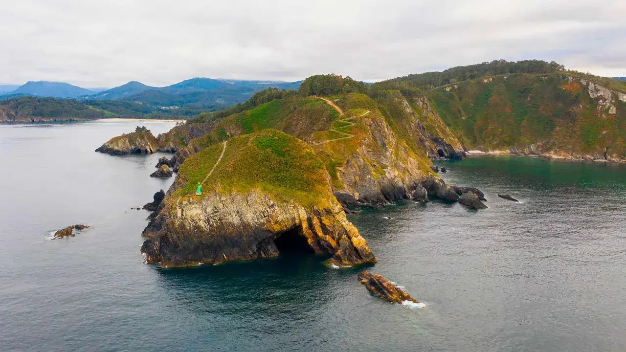 Costa rocosa con una cueva que parece el fuciño do porco. Senderos serpentean por las colinas verdes y un pequeño faro verde vigila el mar.