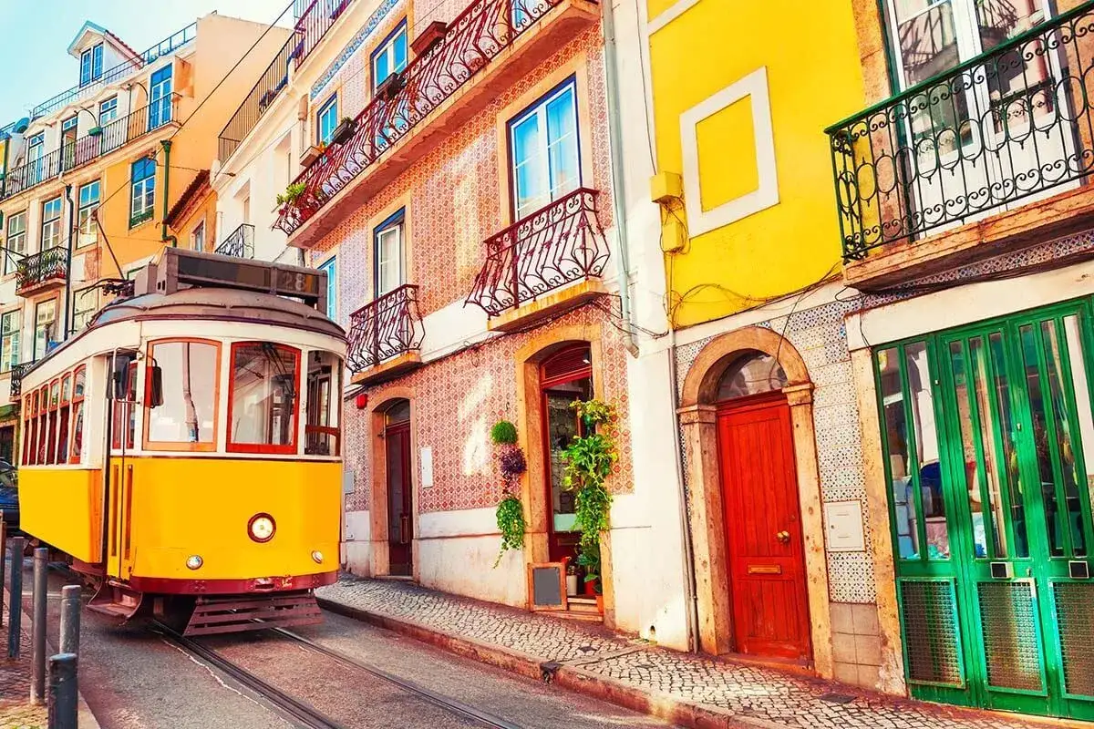 A classic yellow tram rolls down a cobblestone street in Lisbon, past colorful buildings with ornate balconies. Perfect for a day trip!