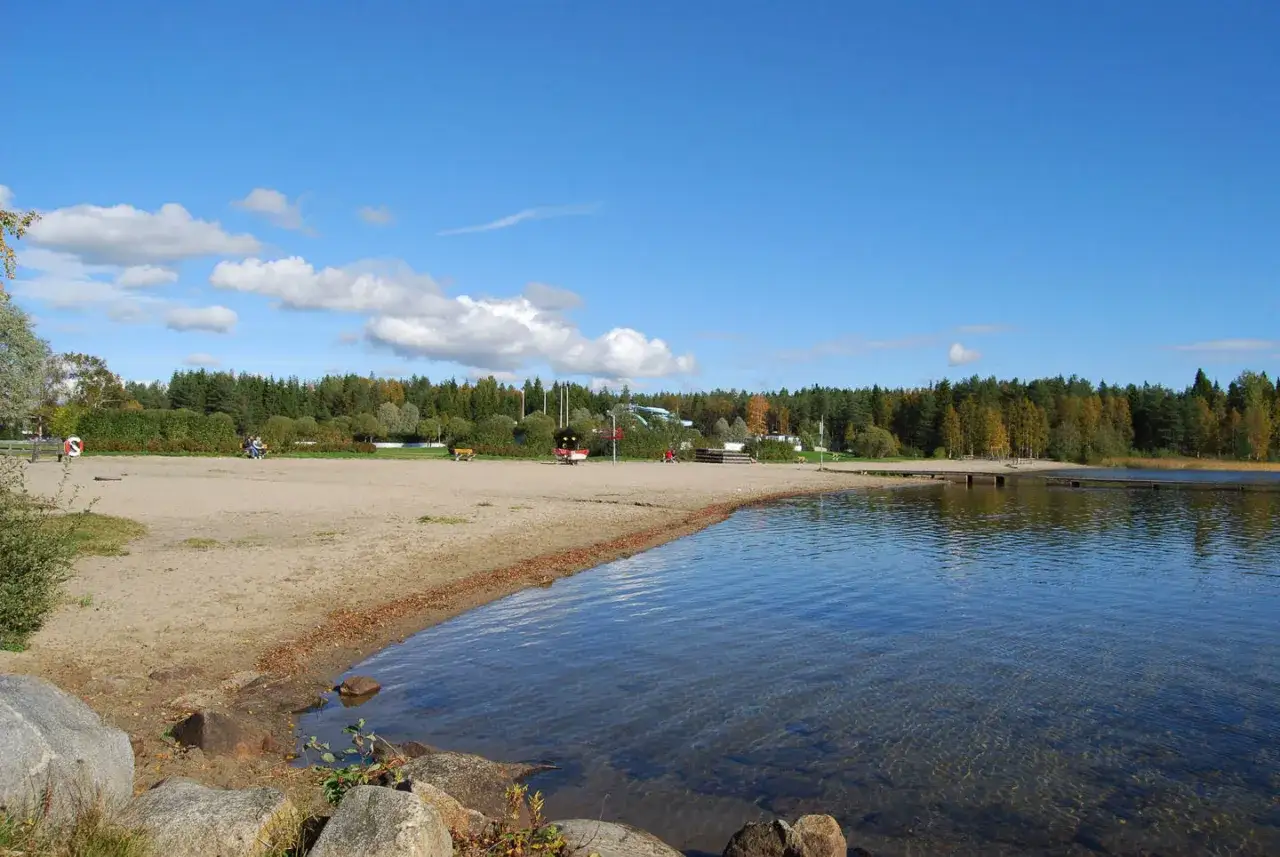 Solig strand vid Nydala camping Ume&aring;. Vatten, sand och skog under en bl&aring; himmel med moln.
