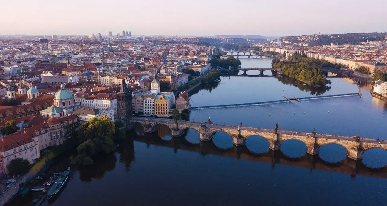 Vue aérienne de Prague, avec le pont Charles et ses arches emblématiques traversant la Vltava. La ville s'étend à perte de vue, un panorama de toits rouges et de dômes.