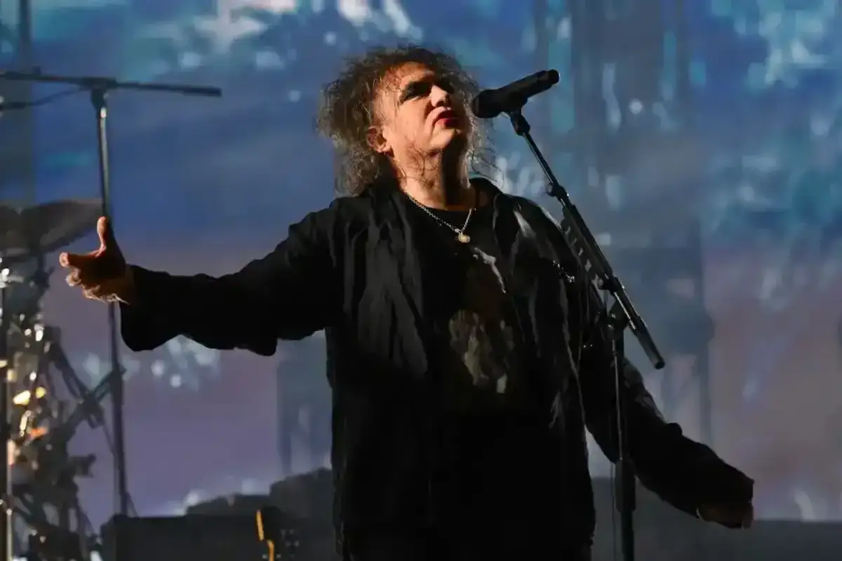 Robert Smith z The Cure śpiewa na koncercie, jego ręka wyciągnięta w geście, jakby zapraszał do wspólnego przeżywania muzyki zespołu The Cure.