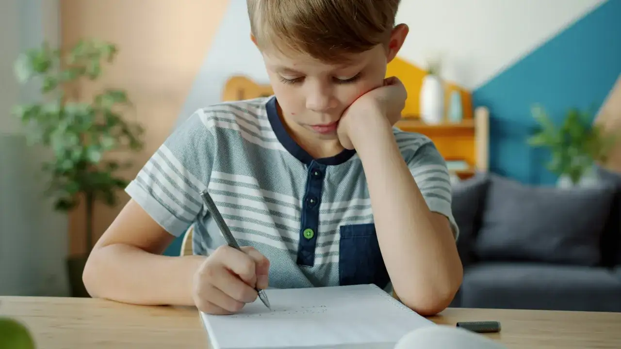 Ein Junge mit Sommersprossen macht konzentriert seine Hausaufgaben. Er stützt den Kopf auf die Hand und schreibt mit einem Stift in sein Heft.