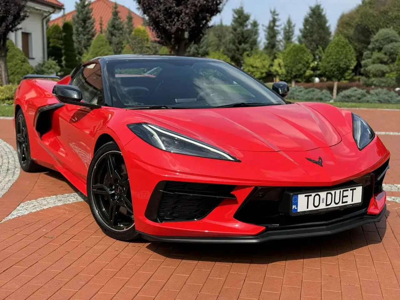 Czerwony Chevrolet Corvette C8 z czarnym dachem i felgami, zaparkowany na podjeździe z czerwonej kostki brukowej, otoczony zielenią.
