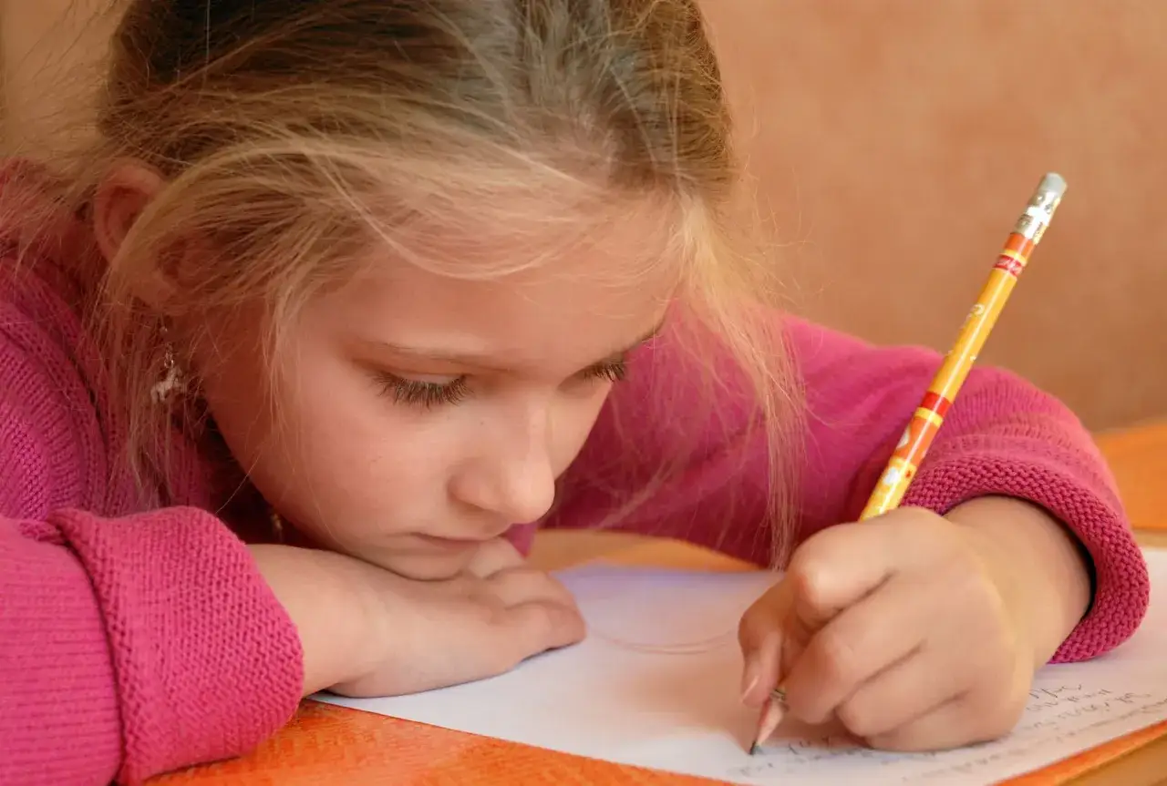 Ni&ntilde;a rubia con su&eacute;ter rosa practica c&oacute;mo mejorar la letra de un ni&ntilde;o, concentrada en su cuaderno.