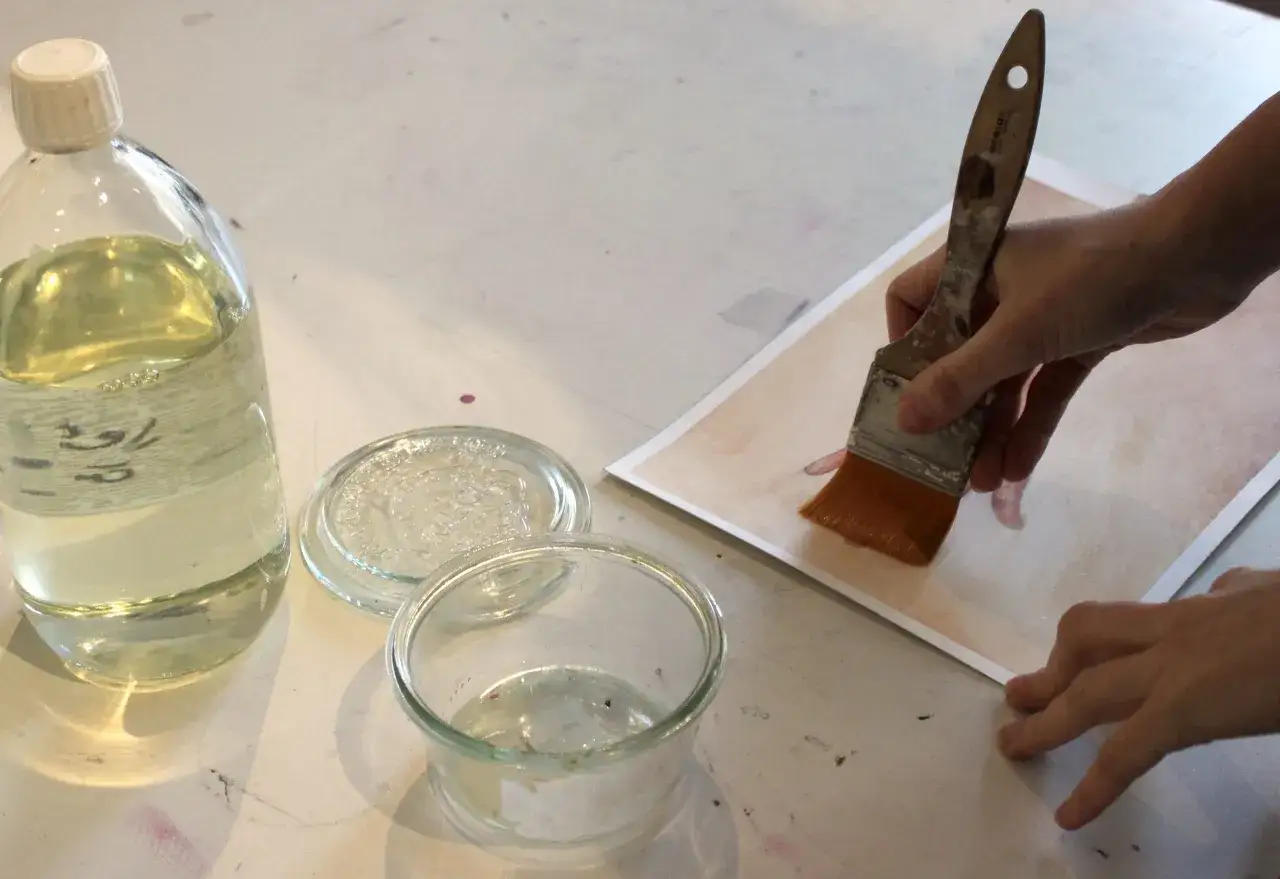 Application d'un vernis sur une œuvre. Une main tient un pinceau imbibé de vernis, prêt à recouvrir le tableau. Une bouteille de vernis et un petit pot sont à proximité.