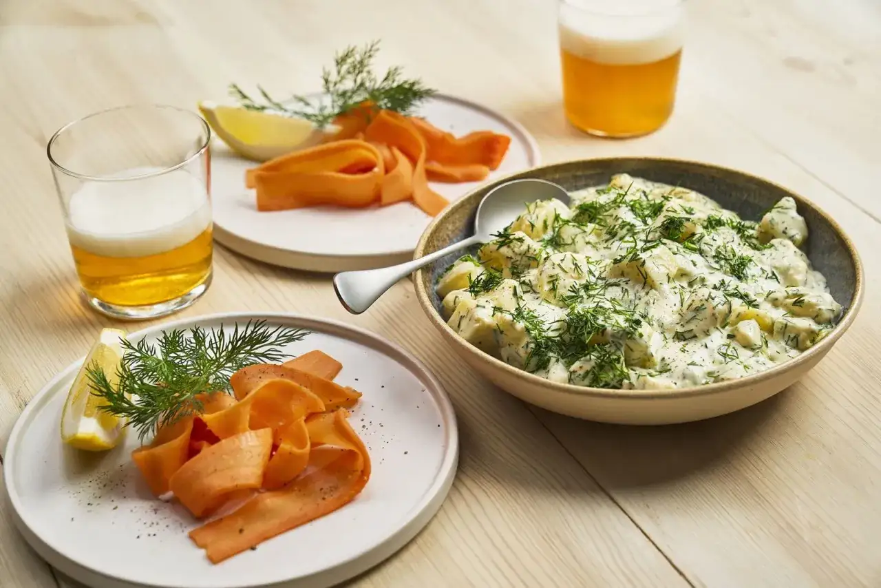 En skål med krämig dillstuvad potatis, serverad med tunna morötter, citronklyftor och öl. En klassisk svensk sommarmåltid.