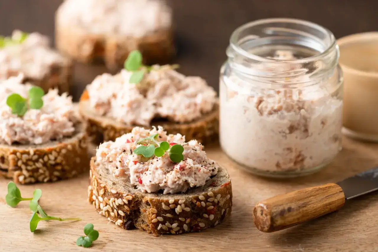 Tartines de pain de seigle garnies de rillettes de thon, décorées de pousses vertes. Un pot de rillettes et un couteau sont à proximité.