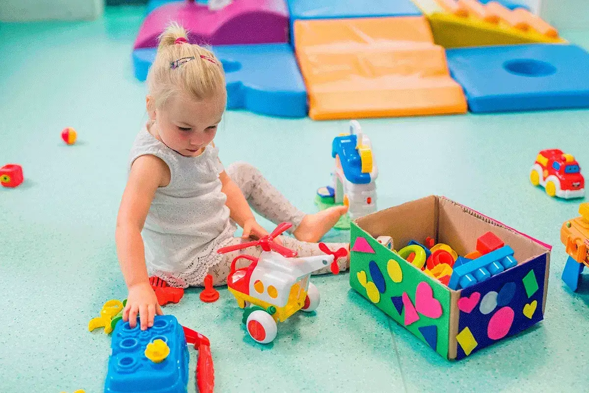 Niña jugando con juguetes, desarrollando su motricidad gruesa. Un helicóptero, un coche y bloques de construcción la entretienen.