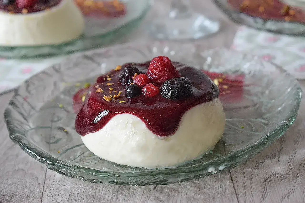 Deliciosa panna cotta con salsa de frutos rojos y pistachos picados, servida en un plato de cristal.