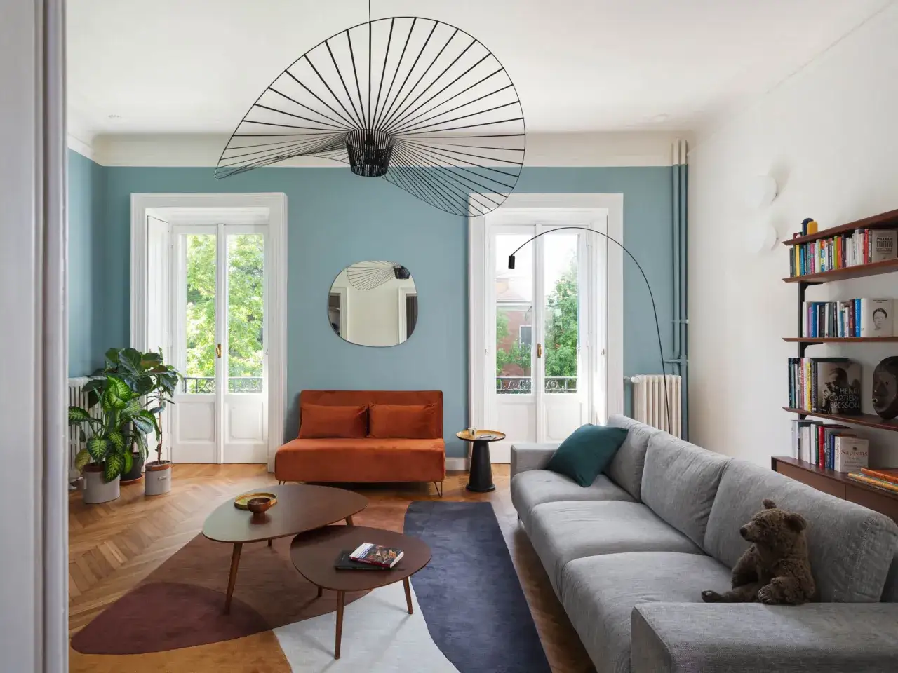 Soggiorno con pareti colorate moderne, divano grigio, arancione e tavolini in legno. Lampadario a raggiera e piante verdi.
